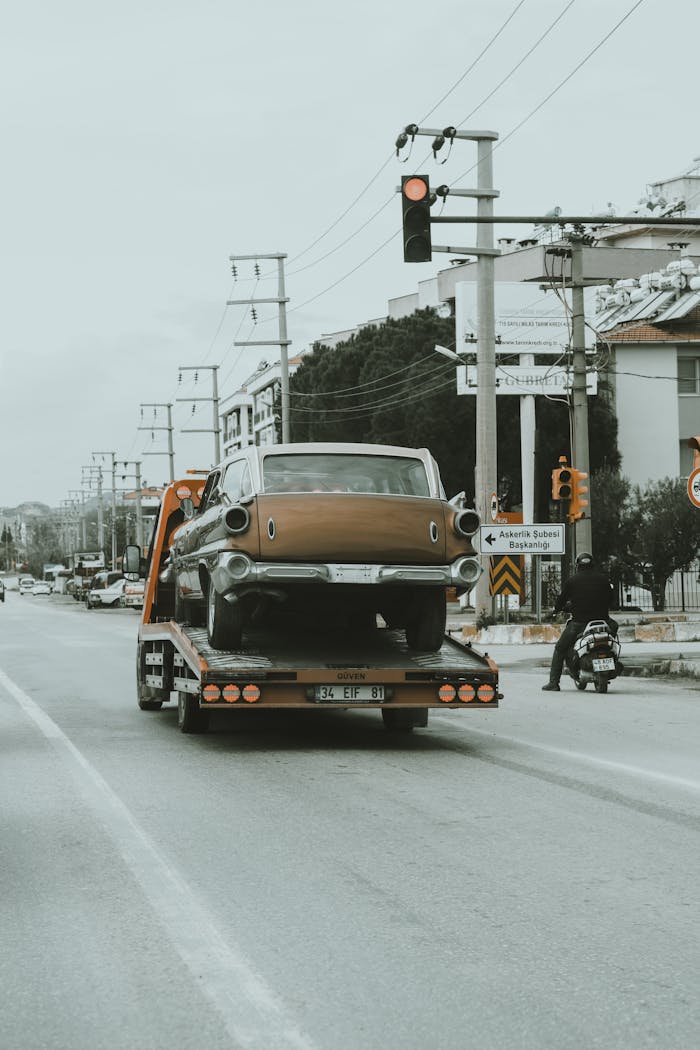 Services-02 A vintage car is being towed through city streets on a tow truck, showcasing an urban environment.